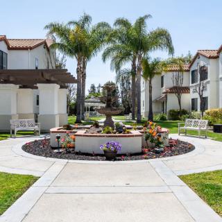 A picturesque residential area with apartments flanking a central walkway and fountain, surrounded by lush palm trees.