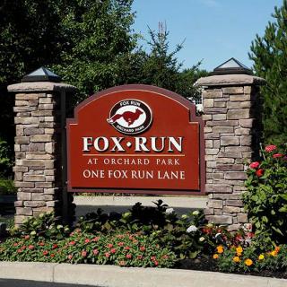Entrance sign for Fox Run at Orchard Park with stone pillars and landscaped flowers.