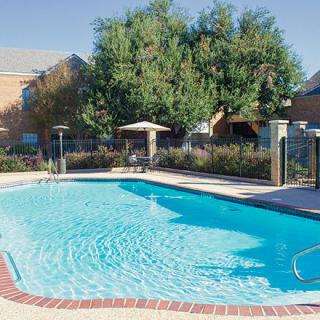 A clear outdoor swimming pool surrounded by trees and residential buildings on a sunny day.