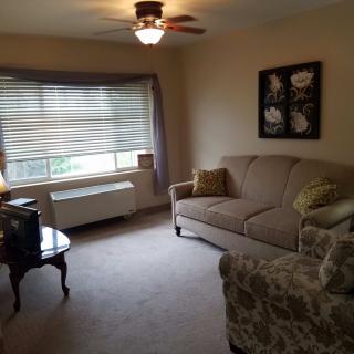 A cozy living room with a sofa, armchair, and a coffee table. Sunlight streams through a window with curtains, and the ceiling fan is on.