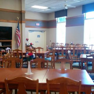 An empty dining room with wooden tables and chairs, featuring an American flag and large windows.