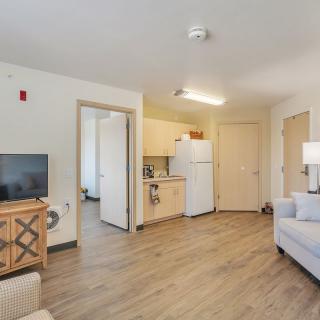 A modern apartment living area with an open kitchen, featuring a sofa, TV, and refrigerator.