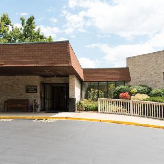 The image shows the entrance of a building with landscaped surroundings and a parking lot under a partly cloudy sky.