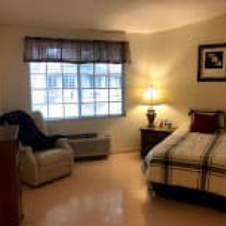 A cozy bedroom with a neatly made bed, an armchair by the window, and a lamp casting a warm glow.