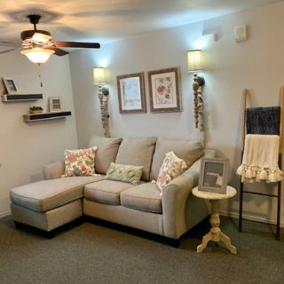 A cozy living room with a sectional sofa, decorated with pillows and wall art, featuring a ceiling fan and ambient lighting.