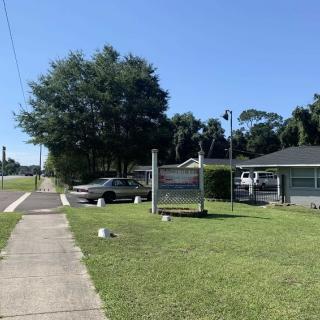 A sunny day with a sidewalk leading past a parked car and building, surrounded by grass and trees.