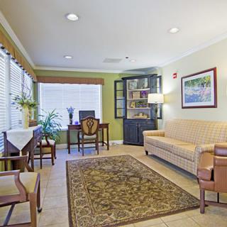 A cozy living room with a sofa, chairs, and a rug, featuring a desk near the window and a bookshelf.