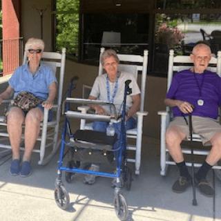Three elderly individuals sitting outside in rocking chairs with a walker and cane nearby.