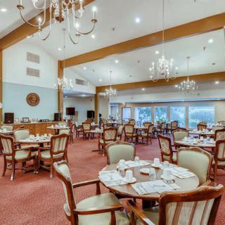 A spacious and elegant dining room with multiple round tables and wooden chairs, illuminated by chandeliers.