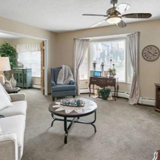 A cozy living room with a sofa, armchair, and coffee table, featuring large windows with curtains and a ceiling fan.