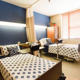 A shared room with two beds in a healthcare facility, featuring patterned bedding and a window with natural light.