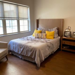 A cozy and well-lit bedroom featuring a neatly made bed with colorful pillows, a comfortable chair, and a nightstand adorned with personal photographs.