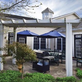 A cozy outdoor patio with a pergola, featuring blue umbrellas, seating, and a grill.