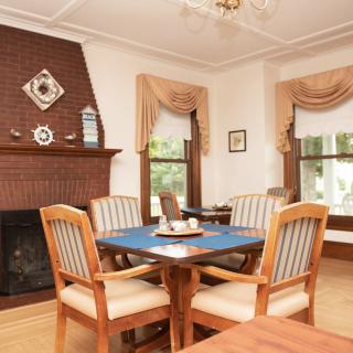 A cozy dining room with a wooden table and upholstered chairs, featuring a brick fireplace and large windows with elegant curtains.