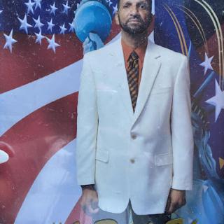 A man dressed in a stylish white suit stands against a vibrant background featuring the American flag.