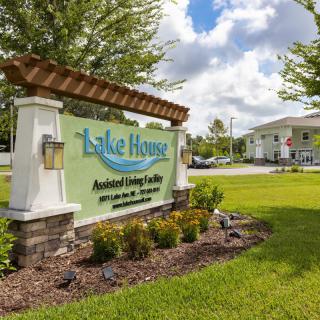 A sign for Lake House Assisted Living Center surrounded by greenery and buildings.