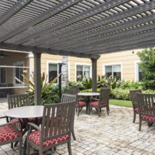 A cozy outdoor patio area with tables and chairs under a pergola, surrounded by well-maintained landscaping.