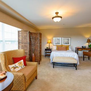 A cozy bedroom with warm lighting, featuring a comfortable armchair, a bed with decorative pillows, and elegant decor.