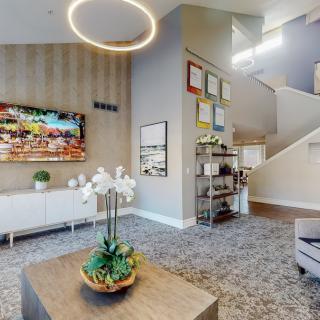 A modern living room with stylish decor, featuring a large chandelier, artwork, and a staircase leading to the upper floor.