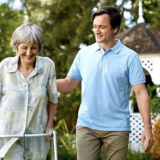 A person assists an elderly woman using a walker in a garden setting.
