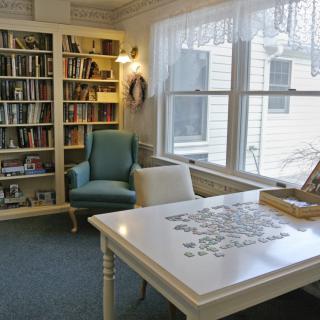 A cozy library corner with a bookshelf, armchair, and a table set up for puzzle-solving near a window.