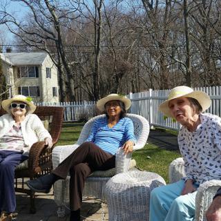 Three elderly women enjoy a sunny day outdoors, dressed in hats and relaxed in their chairs.