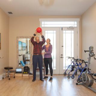 A fitness instructor guides a senior individual in a rehabilitation exercise using a medicine ball in a bright indoor setting.