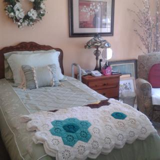 A cozy bedroom with a single bed adorned with a crochet blanket and decorative pillows, accompanied by a nightstand, lamp, and framed art on the wall.