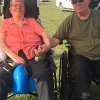 An elderly couple sitting outdoors, holding hands while seated in wheelchairs.