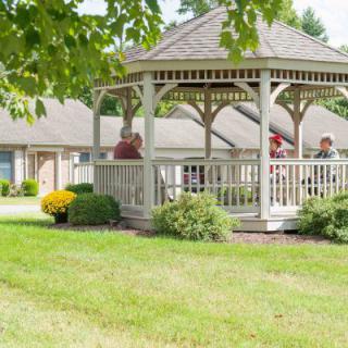 A serene garden scene featuring a wooden gazebo with people sitting inside, surrounded by a lush green lawn and trees.