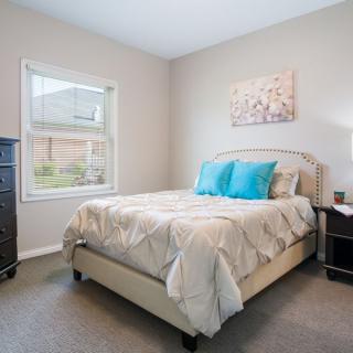 A cozy bedroom with a neatly made bed, two blue pillows, and a nightstand beside it. A window provides natural light, and artwork hangs on the wall above the bed.