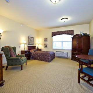 A spacious and well-furnished hotel room featuring a bed, armchair, wooden furniture, and ample lighting.