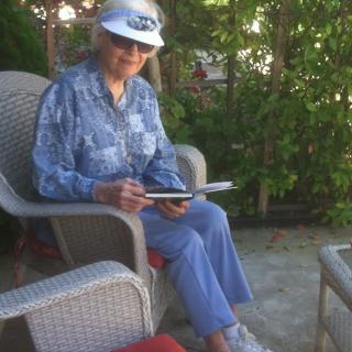 A person sitting outdoors in a chair, wearing a hat and glasses, reading.