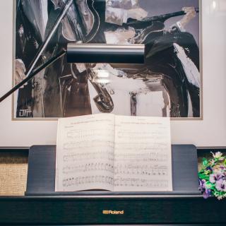 A piano with an open sheet of music, illuminated by a lamp, with artwork in the background.