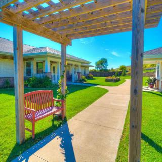A sunny outdoor garden with a pathway, bench, and a pergola leading to residential buildings.
