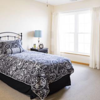 A cozy bedroom with a single bed dressed in patterned bedding, accompanied by a nightstand and lamp near a large window.