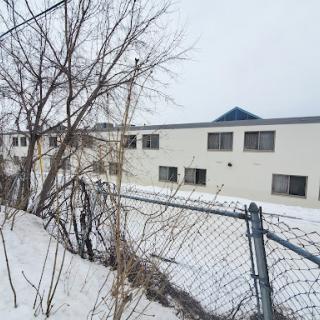 A view of a snow-covered building surrounded by a fence and bare trees on a winter day.