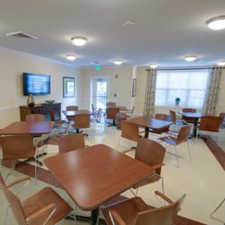 A community dining area with multiple tables and chairs, featuring a television on the wall and large windows with curtains.
