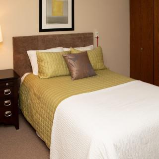 A cozy bedroom with a neatly made bed, featuring green bedding and decorative pillows, alongside a lamp and artwork on the wall.