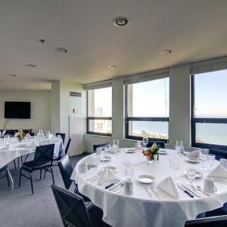 A well-lit dining room with round tables set for an event, offering a stunning ocean view through large windows.