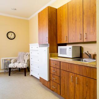 A cozy kitchen corner with wooden cabinets and a microwave on the countertop, featuring a decorative chair and clock against beige walls.
