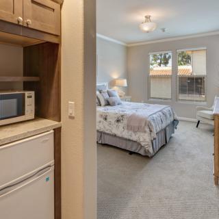 A cozy bedroom with a small kitchenette area, featuring a bed, bedside lamp, microwave, and mini fridge.