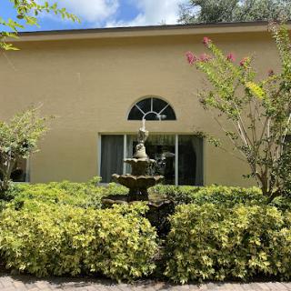 A serene outdoor scene featuring a fountain surrounded by lush greenery in front of a yellow building.
