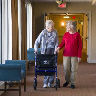Two elderly individuals walking in a hallway, one using a walker and the other providing support.