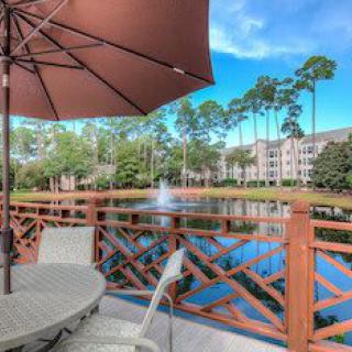 A serene patio with outdoor seating overlooking a lake and fountain surrounded by trees and apartments.