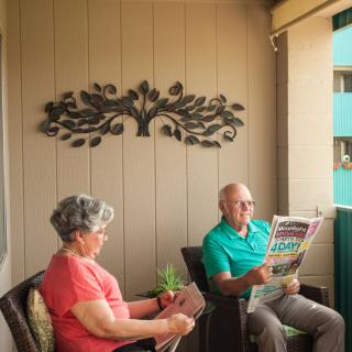 An elderly couple sitting on a balcony, one reading a book and the other a newspaper.