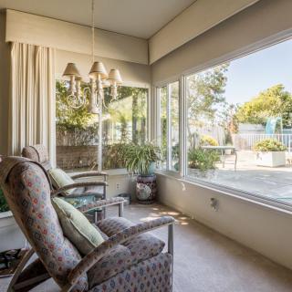 A cozy living room with large windows overlooking a garden, featuring comfortable chairs and indoor plants.