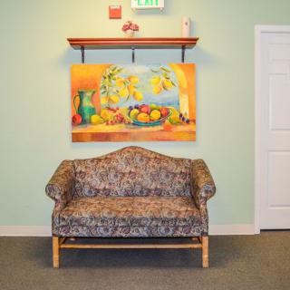 A patterned sofa sits beneath a colorful still-life painting in a well-lit room.