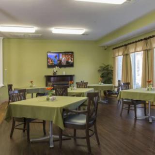 A cozy dining area with green walls, neatly arranged tables and chairs, a television on the wall, and soft lighting.
