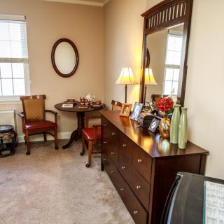 A cozy bedroom with a wooden dresser and matching furniture, including chairs and a small table, decorated with lamps and accessories.